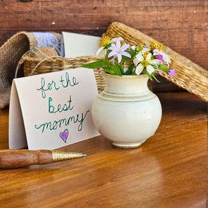 May include: A white card with the text "for the best mommy" written in green and purple ink. A small purple heart is drawn below the text. A white ceramic vase with a bouquet of pink and white flowers sits on a wooden surface.