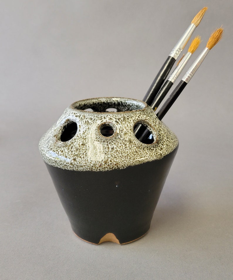 A black and white speckled gloss finish cone shaped ceramic jar that tapers back in at the top and has cut out circle slots for storing painting implements and tools.