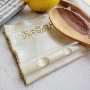 May include: Cream-colored ceramic spoon rest with a decorative swirl design. The rectangular rest has a slightly wavy edge and two circular indentations. A wooden spoon rests on the side, with a lemon and striped cloth in the background.