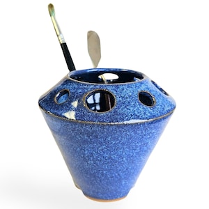 May include: A blue ceramic brush holder with multiple holes for holding brushes and other art supplies. The holder has a speckled blue glaze and a rounded, cone-shaped design.