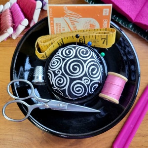 May include: A black sewing pincushion with a white swirl pattern, a yellow tape measure, a pink spool of thread, a thimble, a pair of silver scissors, and a pink seam ripper on a black plate.