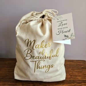 May include: A beige drawstring bag with gold lettering that reads "Maker of Beautiful Things." A small card attached reads "Given with Love Made with Heart" with leaf accents. The bag is on a wooden surface.