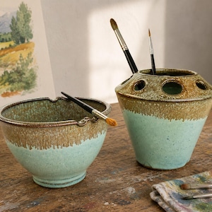 Può includere: Una ciotola in ceramica e un portapennelli abbinato di forma conica con diverse aperture sulla parte superiore. Entrambi i pezzi presentano uno smalto bicolore con una base verde menta maculata e un bordo marrone testurizzato. Sono inclusi due pennelli.