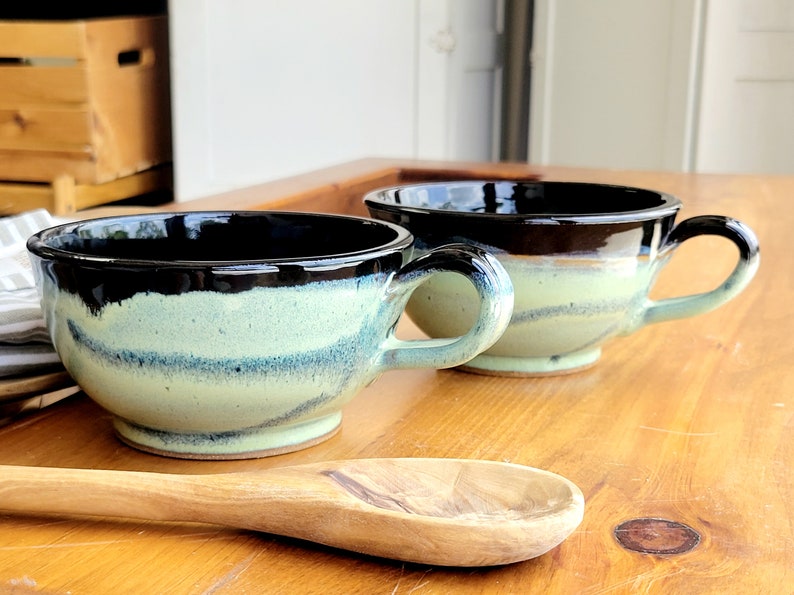 May include: Two handmade ceramic soup bowls with black and green glaze. The bowls have a wide, rounded shape and a single handle. The bowls are sitting on a wooden surface.