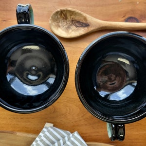 May include: Two black ceramic mugs with green handles. The mugs are empty and have a glossy finish.