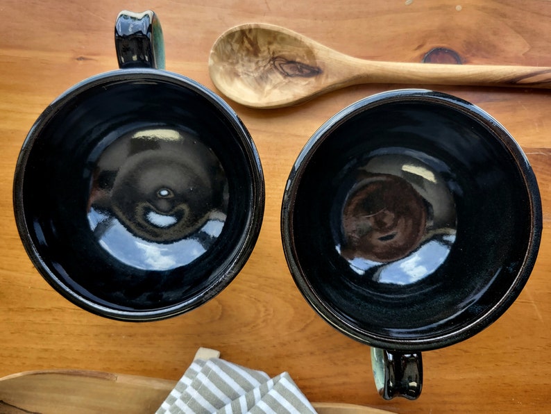 May include: Two black ceramic mugs with green handles. The mugs are empty and have a glossy finish.