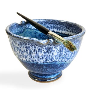 May include: A blue and white ceramic bowl with a paintbrush resting inside. The bowl has a speckled glaze and a wavy rim.