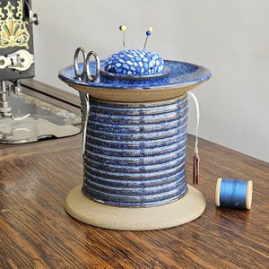 May include: A blue and brown ceramic pincushion and thread holder with a small spool of blue thread. The pincushion has a blue and white patterned top and a small dish for holding sewing supplies.