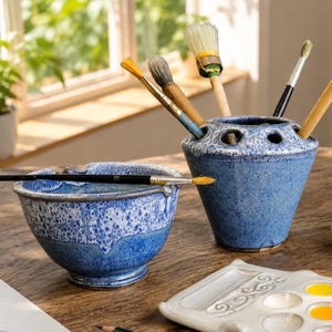 May include: A blue and white speckled ceramic bowl and a matching brush holder with multiple circular openings. Several paintbrushes with wooden handles and bristles are inserted into the holder, while one brush rests across the bowl.