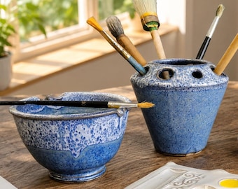 May include: A blue and white speckled ceramic bowl and a matching brush holder with multiple circular openings. Several paintbrushes with wooden handles and bristles are inserted into the holder, while one brush rests across the bowl.