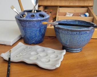 May include: A white ceramic paint palette with nine wells, a blue ceramic bowl, and a blue ceramic brush holder with a brush inside. The items are on a wooden surface.