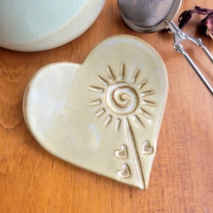 May include: A heart-shaped ceramic tea bag holder with a sun design and three small hearts. The holder is a light beige color with a white glaze.