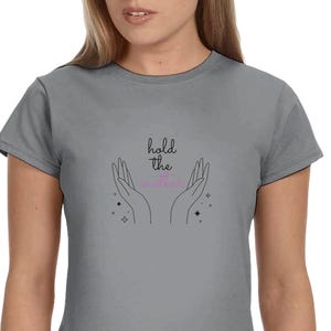 Può includere: Una maglietta grigia con la scritta "hold the" sopra un paio di mani e la parola "magic" in rosa. Il design include anche piccole grafiche a stella e puntini.