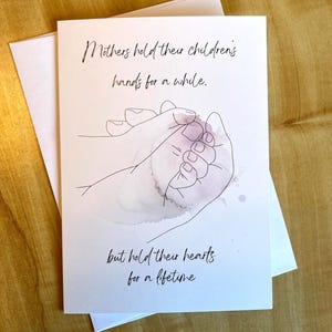 Può includere: Biglietto di auguri bianco con un disegno a tratto di due mani, una adulta e una di bambino, con effetto acquerello. Il biglietto recita: "Le madri tengono le mani dei loro figli per un po', ma tengono i loro cuori per tutta la vita."