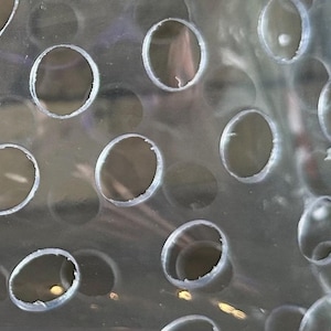 May include: Close-up of a transparent plastic sheet with numerous circular holes. The holes are evenly spaced and vary slightly in size. The edges of the holes have a slightly rough texture, and the overall appearance is clean and modern.