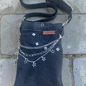 May include: A black denim crossbody bag featuring a black adjustable strap, silver pyramid studs, and a layered silver chain attachment with star charms. A brown leather-like rectangular patch with the text CAMPELLI is sewn on the front.