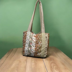 May include: A patchwork tote bag with floral and striped patterns in brown, green, and white. The bag has long, striped handles and a small, rectangular leather tag. The bag is made of quilted fabric.