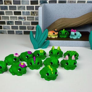 May include: A collection of small, green frog figurines with various accessories, including flowers and horns. Some frogs are yellow and blue. The figurines are arranged on a white surface with a decorative backdrop and green plant accents.