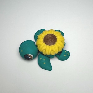May include: A whimsical teal turtle figurine with a bright yellow sunflower on its back. The turtle has a detailed eye and yellow spots on its flippers. The sunflower has a brown center and yellow petals.