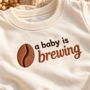 May include: An off-white crewneck sweatshirt featuring brown embroidery of a coffee bean and the text "a baby is brewing" centered on the chest. The garment has a ribbed collar and a soft, smooth fabric texture throughout the front panel.