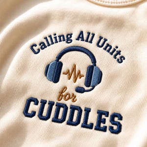 Op de afbeelding: Een crèmekleurig kledingstuk met een geborduurd ontwerp van een blauwe koptelefoon met een goudkleurig geluidsgolfpictogram in het midden. De tekst Calling All Units for CUDDLES is in donkerblauw en goudkleurig garen geborduurd.