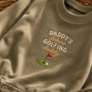 May include: An olive green long-sleeved bodysuit featuring embroidered text that reads DADDY'S future GOLFING buddy in white and yellow thread. Below the text is an embroidered green golf putting green with a red flag and a white golf ball.
