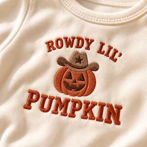 May include: An off-white long-sleeved shirt featuring an embroidered graphic of an orange jack-o'-lantern wearing a brown cowboy hat. The text "ROWDY LIL' PUMPKIN" is embroidered in brown block lettering above and below the pumpkin graphic.