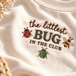 Könnte beinhalten: Ein cremefarbenes, geripptes Sweatshirt mit braunem Sticktext, der the littlest BUG IN THE CLUB lautet. Das Design umfasst einen roten Marienkäfer, einen grünen Käfer sowie eine gelb-braune Biene, die um den Text herum gestickt sind.