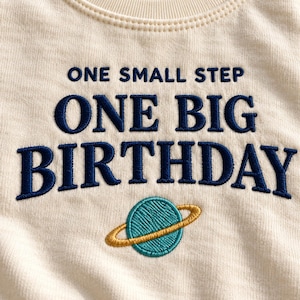 Może przedstawiać: Kremowy t-shirt z ciemnoniebieskim haftem o treści ONE SMALL STEP ONE BIG BIRTHDAY. Pod tekstem znajduje się wyhaftowana turkusowa planeta ze złotym pierścieniem, umieszczona centralnie na klatce piersiowej odzieży.