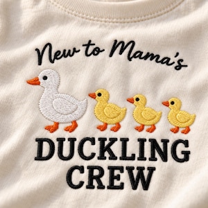 Op de afbeelding: Een off-white shirt met zwarte geborduurde tekst die luidt New to Mama's DUCKLING CREW. Het ontwerp bevat een geborduurde witte volwassen eend met een oranje snavel en poten, gevolgd door drie kleinere gele eendjes met oranje snavels en poten.