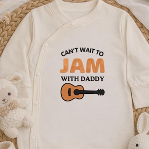 May include: Cream-colored baby onesie with the text "Can't wait to JAM with Daddy" in orange and black. A cartoon guitar graphic is below the text. The onesie is laid on a woven mat with a stuffed lamb and a stuffed bunny.