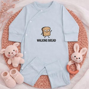 Puede incluir: Mono de bebé azul claro con una imagen de dibujos animados de una rebanada de pan con el texto "WALKING BREAD". La imagen incluye juguetes para bebés: un osito de peluche, zapatos de bebé y un mordedor de conejito.