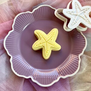 May include: A pale yellow starfish-shaped biscuit with embossed texture details sits on a scalloped mauve plate. Beside the plate, a white plastic starfish biscuit cutter is visible, featuring a matching five-pointed design with small raised dots.