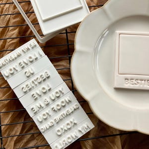 May include: White cookie cutters and stamps for creating cookies with messages. The stamps include phrases like "Bestie", "I love you", and "XOXO". A cookie shaped like a polaroid is on a white plate.