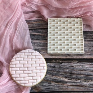 May include: A round biscuit decorated with a brick pattern, next to a square brick-patterned mould. The biscuit has a light pink icing, and the mould is off-white. A pink fabric is draped on a wooden surface.