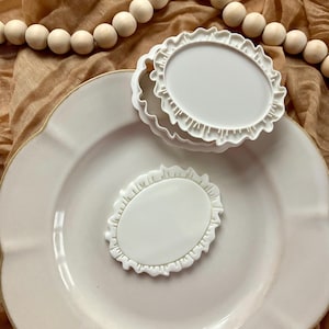 May include: A stack of three white, oval-shaped cookie cutters with a ruffled edge, and one separate cutter, are on a white plate. A string of wooden beads is in the background.