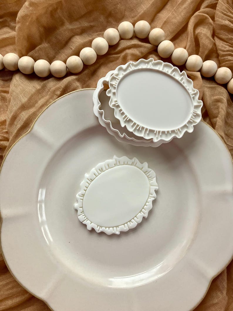 May include: A stack of three white, oval-shaped cookie cutters with a ruffled edge, and one separate cutter, are on a white plate. A string of wooden beads is in the background.