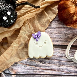 May include: A white ghost-shaped biscuit decorated with a purple bow, smiling face, and a scalloped edge. The biscuit is on a wooden surface, with a decorative spider and pumpkin in the background. A Halloween-themed treat.