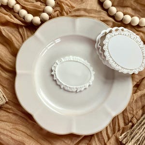 May include: A white scalloped-edge plate with a matching oval-shaped decorative piece. Two additional oval pieces with a lace-like border are stacked nearby. The items are on a textured, tan surface, with wooden beads and twine accents.