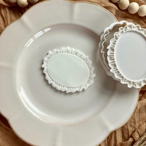 May include: A white ceramic plate with a scalloped edge, a small oval-shaped dish, and two small round dishes with ruffled edges. The items are arranged on a tan surface with wooden beads.