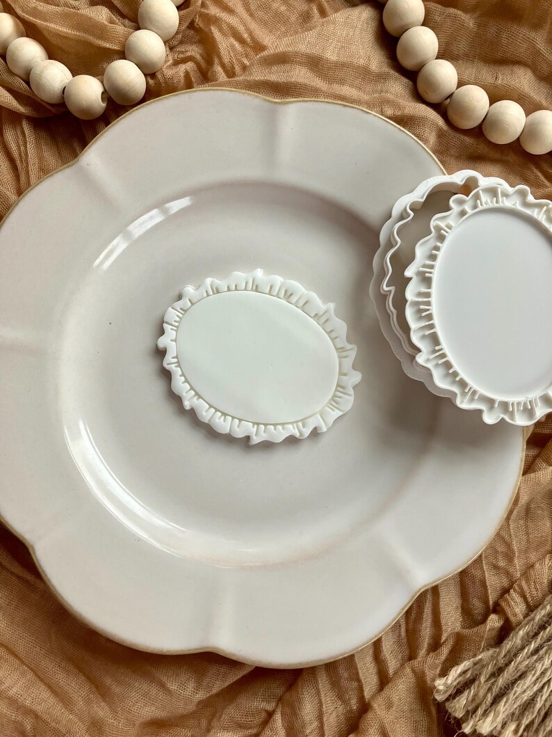 May include: A white ceramic plate with a scalloped edge, a small oval-shaped dish, and two small round dishes with ruffled edges. The items are arranged on a tan surface with wooden beads.