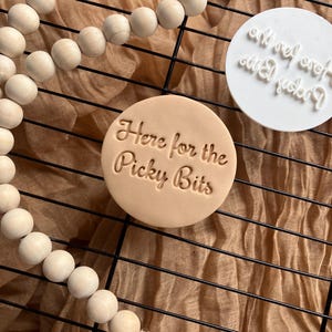 May include: A light tan, circular stamp with the phrase "Here for the Picky Bits" in a cursive font. A white stamp with the text "and yet, even still, I fuckin'" is also visible. The stamps are on a black wire rack with wooden beads.