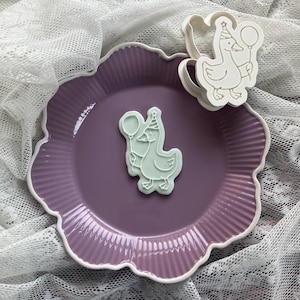 May include: A purple plate with a scalloped edge holds a light green goose-shaped cookie cutter and a matching cookie. The goose wears a party hat and holds a balloon. The plate sits on a white lace cloth.