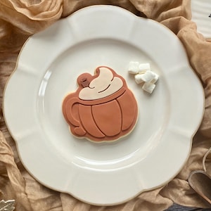 May include: A pumpkin spice latte biscuit with white icing and a cherry, on a white scalloped plate. Several small marshmallows are scattered nearby. The plate rests on a tan fabric.