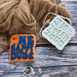 May include: An orange and blue cookie with the words "ONE HAPPY DUDE" and a smiley face. A white silicone mold with the same text and smiley face is next to the cookie. The background includes a tan fabric and a wooden surface.