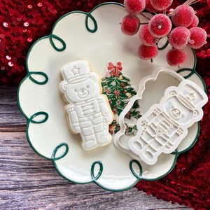 May include: A white plate with a green scalloped edge features a bear-shaped biscuit and a cookie cutter. The biscuit is decorated as a toy soldier. A sprig of red berries sits on the plate, and a Christmas tree is visible.