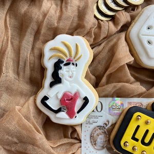 May include: A decorated sugar cookie shaped like a showgirl with a red bodice, black gloves, and a feathered headdress. Other cookies and British currency are also visible.