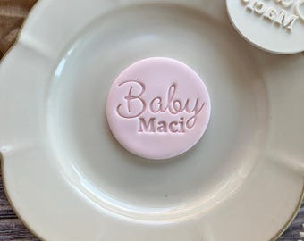 May include: A light pink, circular fondant with the words "Baby Maci" in cursive lettering. A white, circular stamp with the name "Maci" is also visible. The items are on a white, scalloped-edge plate.