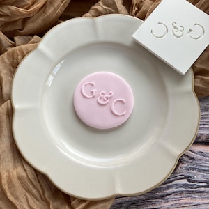 May include: A cream-coloured plate with a scalloped edge displays a pink fondant disc embossed with the letters "G&C". A white square mould with the same letters sits beside the plate. A brown fabric forms the background.