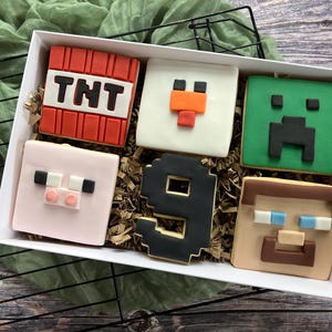 May include: A white box containing six square Minecraft-themed cookies. Designs include a TNT block, a chicken, a Creeper, a pig, the number 9, and a character. The cookies are presented on brown paper. The box is set against a green cloth.